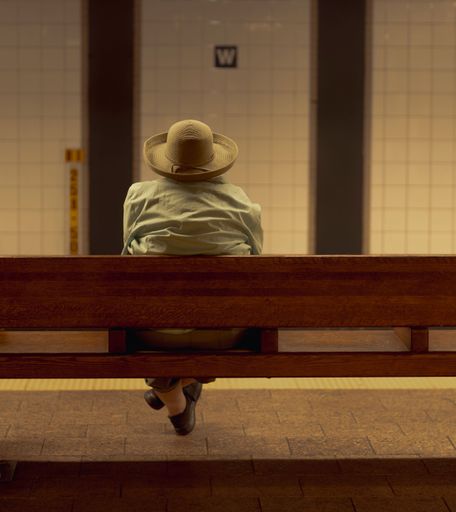 The back of an old woman wearing hat sitting on a bench in subway station in New York City, with white tiled wall in front of her