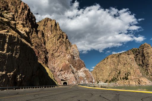 A scenic view of a mountain road leading to a tunnel with a clear blue sky and clouds.