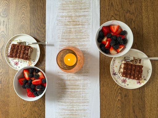 A top view of a cozy dinner table featuring a wooden table runner, a lit candle, and two plates with slices of a layered dessert on each side.