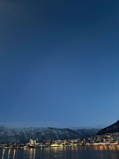 A vertical view of a small village illuminated at night, with snow-covered hills under blue dusk sky in the background in Tromso, Troms county, Norway