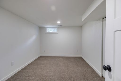 An interior view of a renovated empty room, with carpet flooring and small window on the white wall, illuminated with bright light