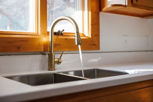 A closeup shot of the modern stainless faucet of a sink in a renovated kitchen illuminated with sunlight through the window in the backdrop
