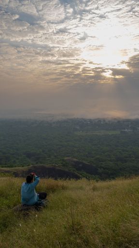 A person sitting on a grassy hilltop, photographing a scenic landscape with a cloudy sky during sunset.