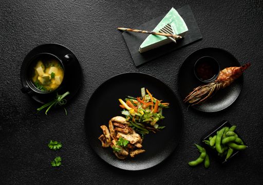 A top view of a Japanese meal with miso soup, grilled chicken, shrimp, edamame, and a slice of green cake on a black textured background.