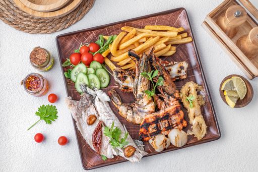 A top view of a seafood platter with grilled shrimp, fish, calamari, fries, and fresh vegetables on a brown plate.