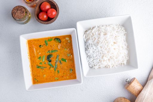 A top view of a delicious curry with vegetables and a side of white rice in square bowls, garnished with cherry tomatoes and spices.
