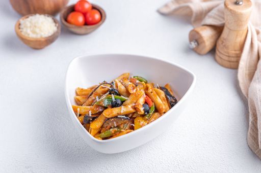 A bowl of penne pasta with vegetables and tomato sauce, garnished with parmesan cheese, on a white table with ingredients in the background.