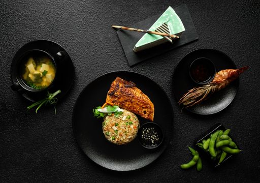 Top view of a Japanese meal, including miso soup, grilled salmon, fried rice, grilled shrimp, and edamame on a textured black background.