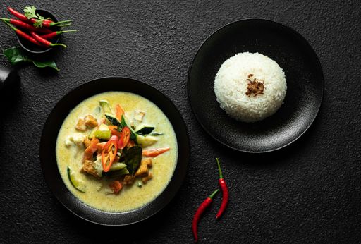 A top view of a plate of Thai green curry with vegetables and tofu served with steamed rice on a black background.