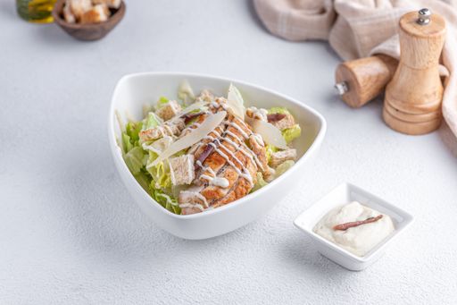 A fresh Caesar salad with grilled chicken, croutons, and Parmesan cheese served in a white bowl with a side of Caesar dressing.