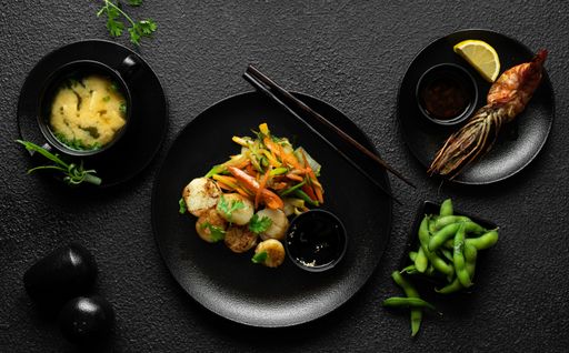 Beautifully arranged Japanese meal with miso soup, grilled shrimp, edamame, and a main dish of scallops and vegetables on a black textured background.