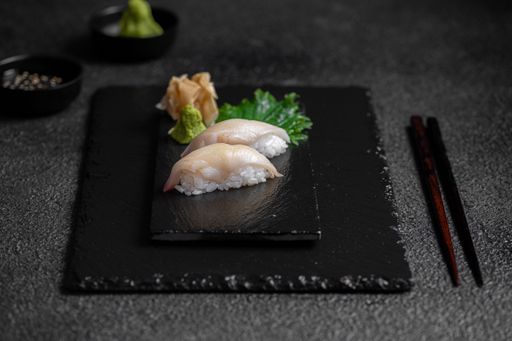 A close-up of two pieces of nigiri sushi on a black slate plate with chopsticks and garnishes.