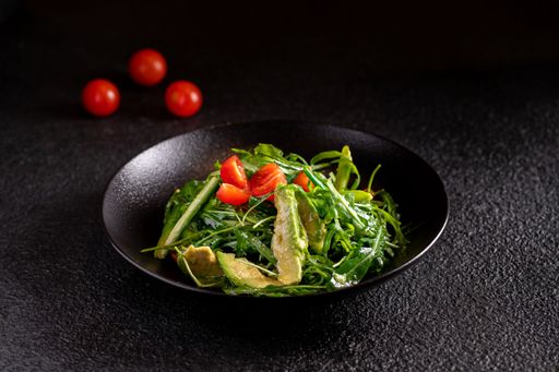 A fresh green salad with avocado and cherry tomatoes served in a black bowl on a dark textured background.