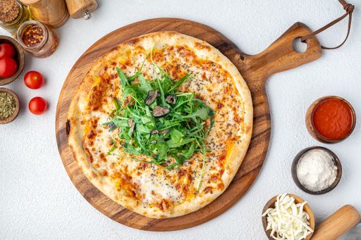 Top view of a freshly baked pizza with arugula and truffle slices on a wooden board, surrounded by ingredients on a white background.
