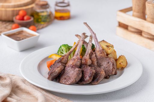A plate of grilled lamb chops with roasted potatoes and mixed vegetables, served with a side of sauce, set on a white table.