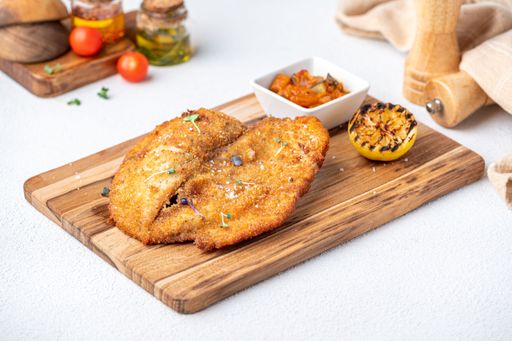 A delicious breaded schnitzel served on a wooden board with grilled lemon and a side of vegetable relish.