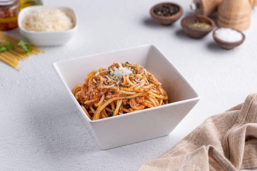 Close-up of a bowl of spaghetti bolognese topped with cheese, with ingredients and spices in the background on a white surface.