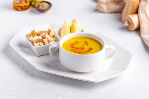 A close-up of a bowl of yellow soup garnished with paprika, served with croutons and lemon wedges on a white plate.