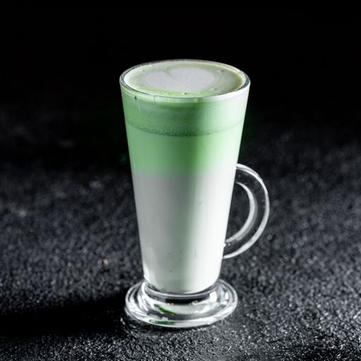 A glass of matcha latte with a heart design on the frothy green top standing on a dark textured surface.