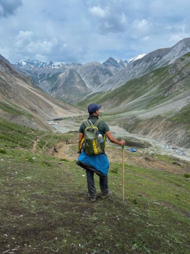 A lone male hiker with a backpack and walking stick looking out over a scenic mountain valley under a cloudy sky.