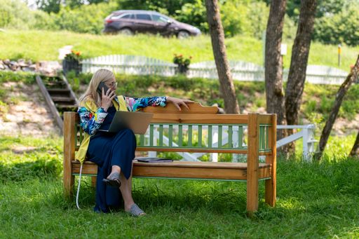A middle-aged woman working on a laptop while talking on the phone, sitting on a bench in a lush park