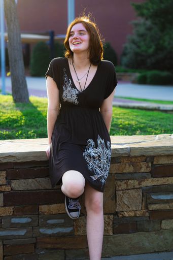 Young woman in a black dress sitting on a stone wall in a park, smiling and enjoying the sunny day.