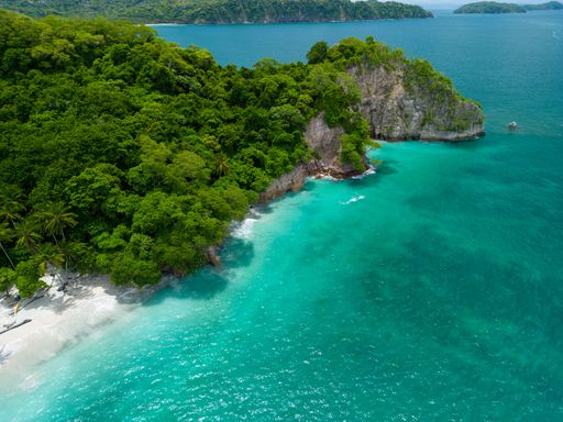 Aerial view of Quesera beach with turquoise waters and lush green forest, featuring a rocky cliff and clear blue sky.