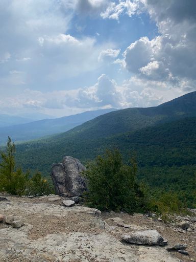 A scenic view of a mountainous landscape under a partly cloudy sky, with rocks and greenery in the foreground.