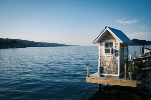 A serene lakeside view with a small wooden shack on a dock, clear blue water, and distant hills under a bright blue sky.
