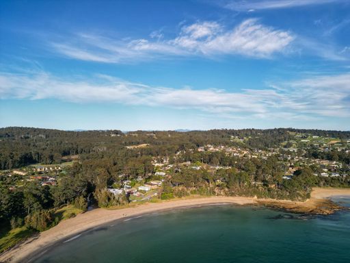 Aerial view of a coastal town with a sandy beach, lush green forest, and a blue sky with scattered clouds.