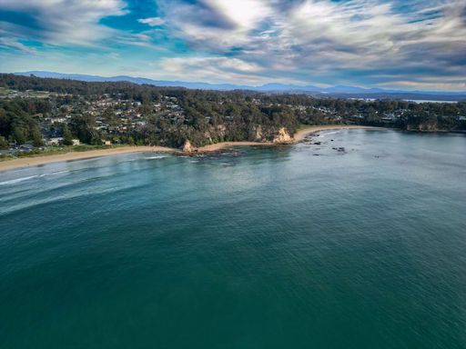 Aerial view of a coastal town with a sandy beach, lush green trees, and distant mountains under a partly cloudy sky.