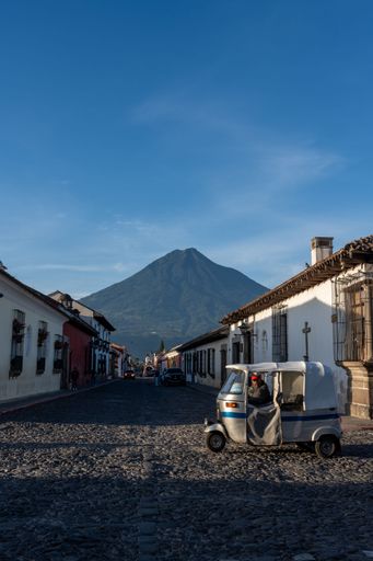 A vertical view of a Tuktuk standing on the street between white houses with mountain background in Antigua Guatemala at sunrise