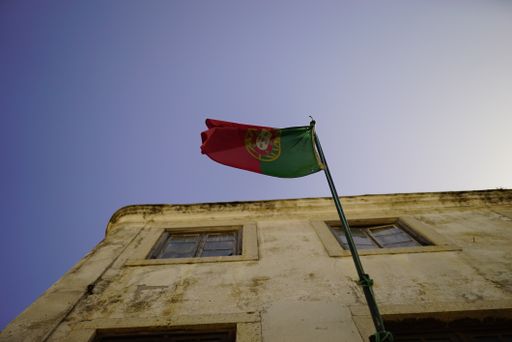 A low-angle shot of the flag of Portugal winding against the blue sky and an old buiding
