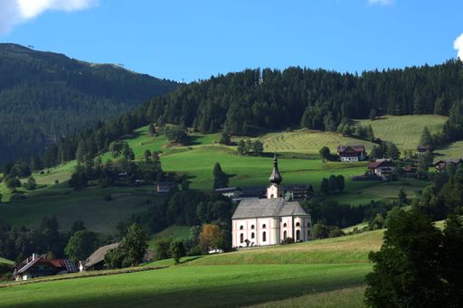 A charming white church with a steeple surrounded by green hills, meadows, and forests under a clear blue sky.