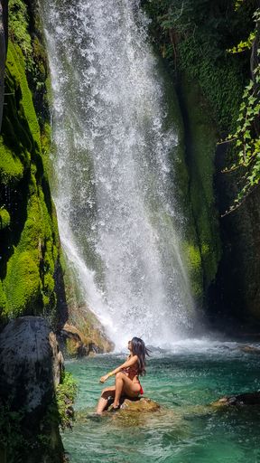 A vertical shot of  a woman sitting on a rock under a waterfall