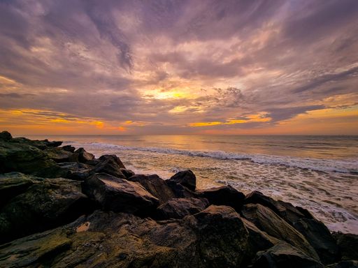 A stunning sunset over a rocky beach with waves crashing against the shore and a dramatic sky filled with colorful clouds.