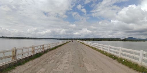 A long, weathered road stretching across a lake under a partly cloudy sky, with distant mountains in the background.
