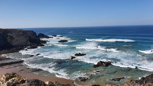 Scenic view of a rocky coastline with waves crashing against the shore under a clear blue sky.