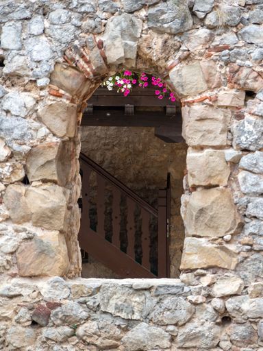 An arched window in an aged stone wall at a medieval fortification. Flowers appears on the top of the window