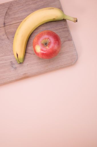 A vertical shot of an apple and banana on a wooden chopping board isolated on light pink background