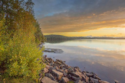 A tranquil lakeside scene with a rocky shore and lush greenery under a cloudy sunset sky.