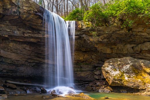 A picturesque waterfall cascading over a rocky cliff into a serene pool below, surrounded by lush greenery and bare trees.