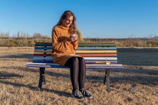 A shot of the Latin woman drinking tea on wooden bench outdoors during an autumn day.