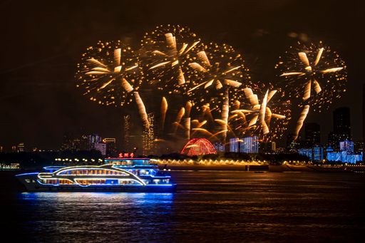 A vibrant night scene with a boat illuminated by colorful lights on the river and fireworks lighting up the sky over a cityscape.