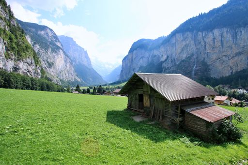 A picturesque view of a rustic barn in a lush green valley surrounded by towering mountains on a clear day in Lauterbrunnen, Switzerland.