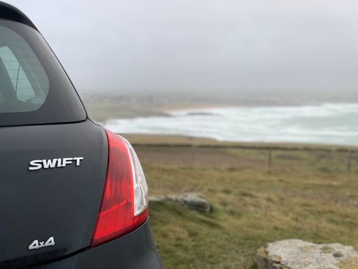 The tail light of a black Suzuki Swift parked on a beach by foam waves and cloudy sky