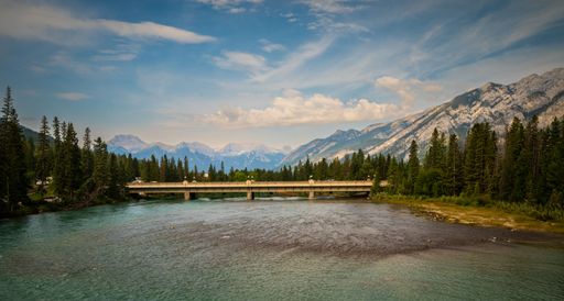 Scenic view of a bridge over a river with mountains and pine trees in the background under a clear blue sky.