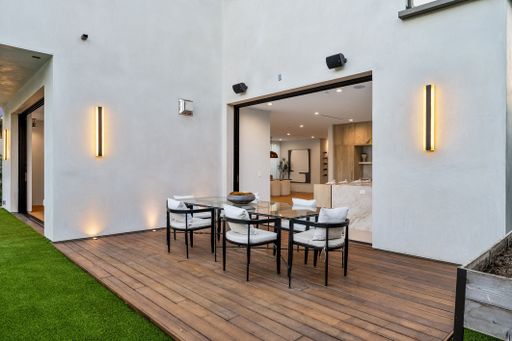 Modern outdoor dining area with a wooden deck, glass table, and chairs, adjacent to a contemporary interior space.