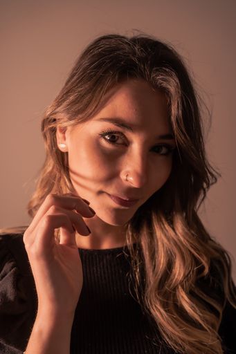 A vertical shot of a young Caucasian woman with long hair posing in front of a brown wall while looking into the camera