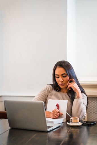 A vertical shot of a smart Hispanic woman writing in her notebook as she works on her laptop